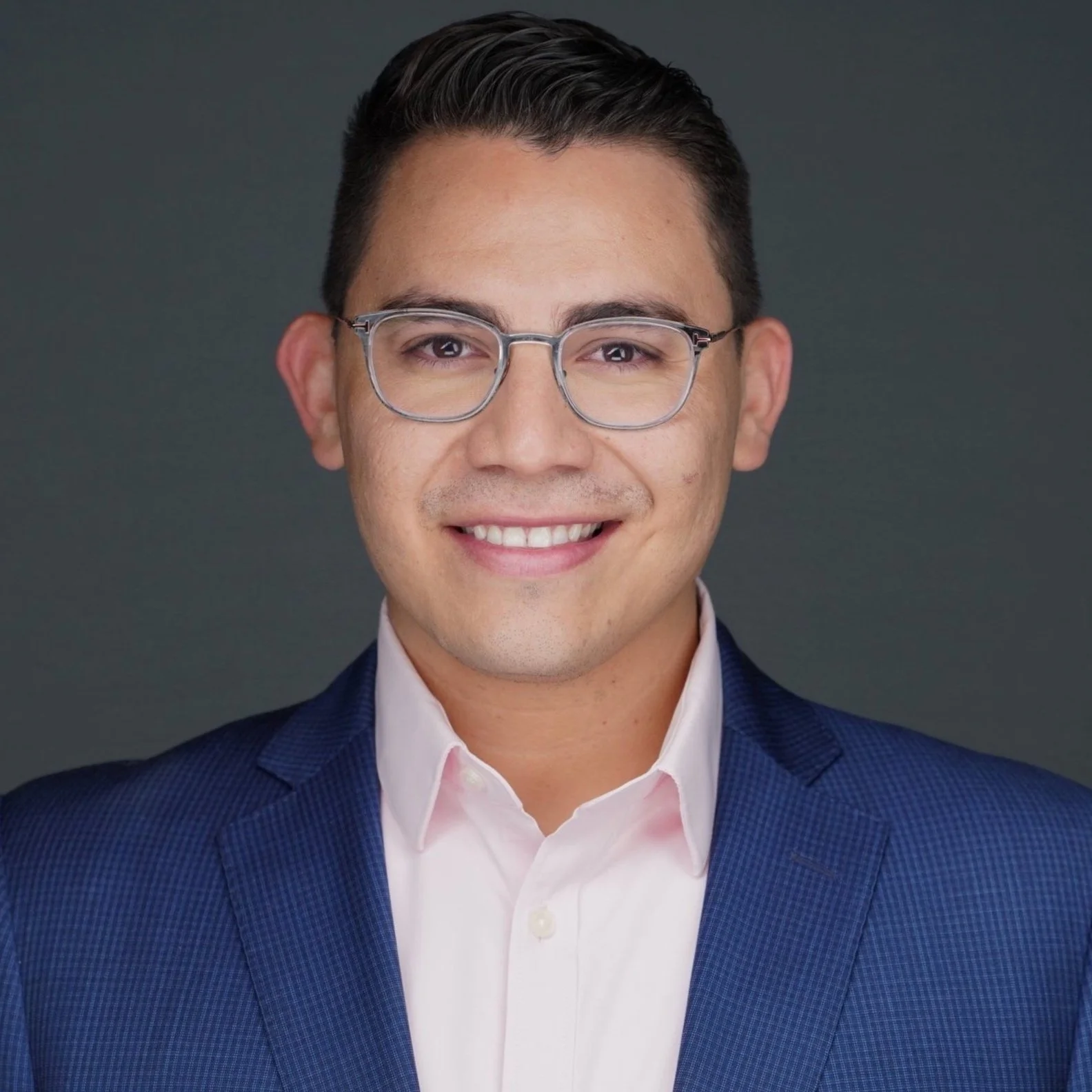 Professional headshot of a smiling man with dark hair, glasses, wearing a navy blue blazer and white shirt, against a dark gray background.