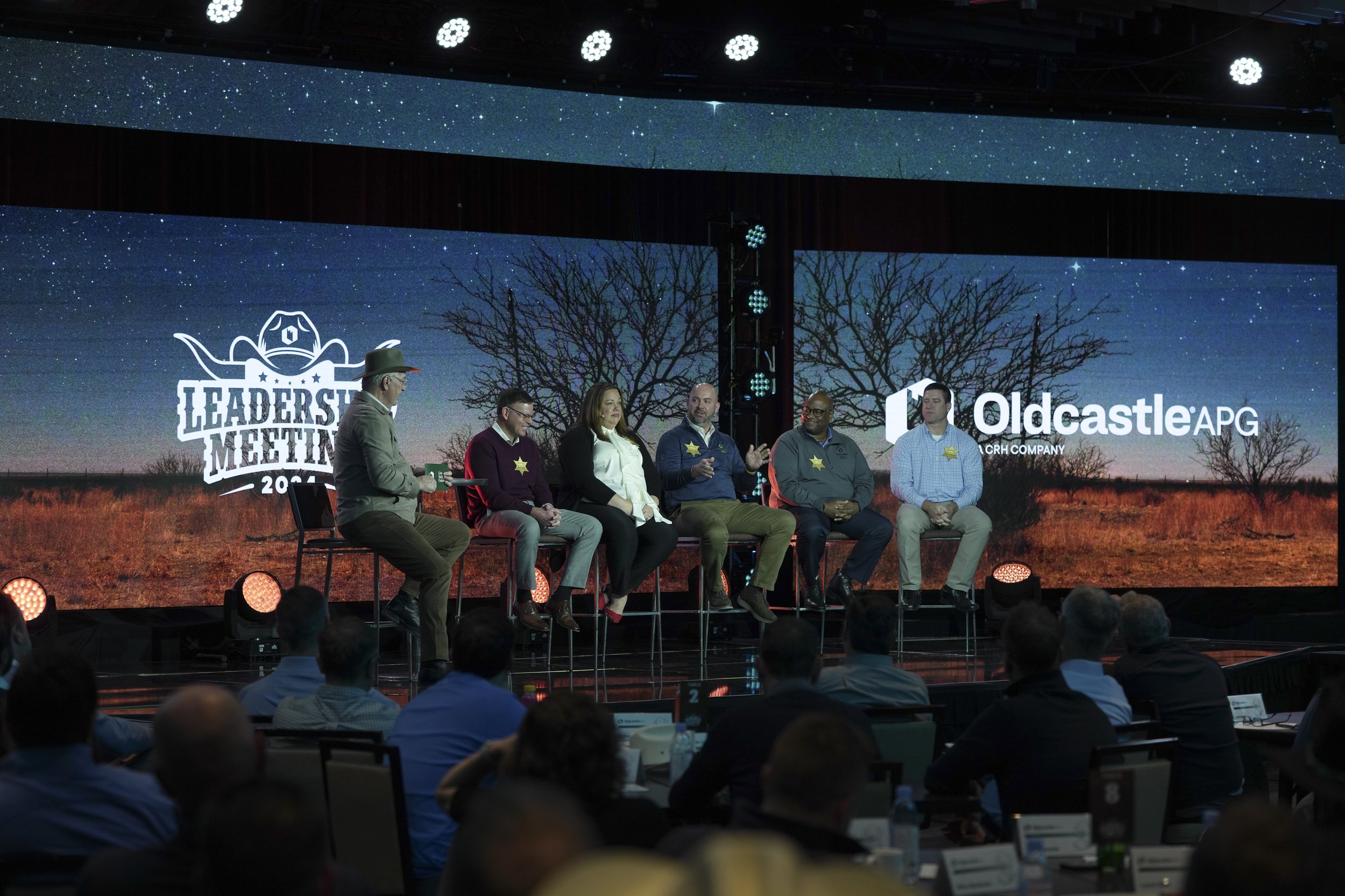Panel discussion at a leadership meeting with six speakers on stage, seated on high stools, with a large screen behind displaying the event logo and a rural landscape background, and an audience in front.