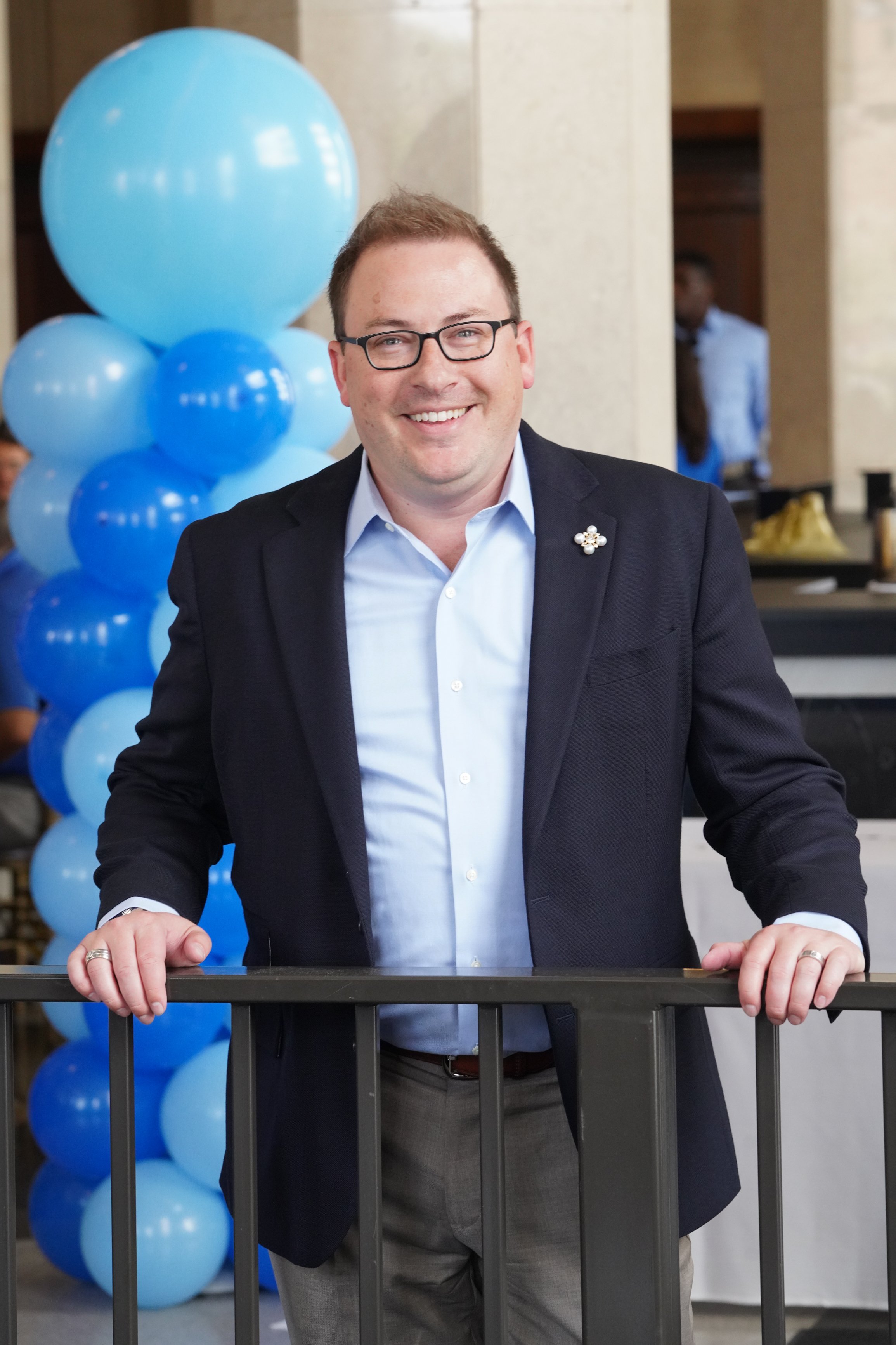 A smiling man dressed in a dark blazer, light blue shirt, and glasses standing behind a railing with blue balloons in the background.