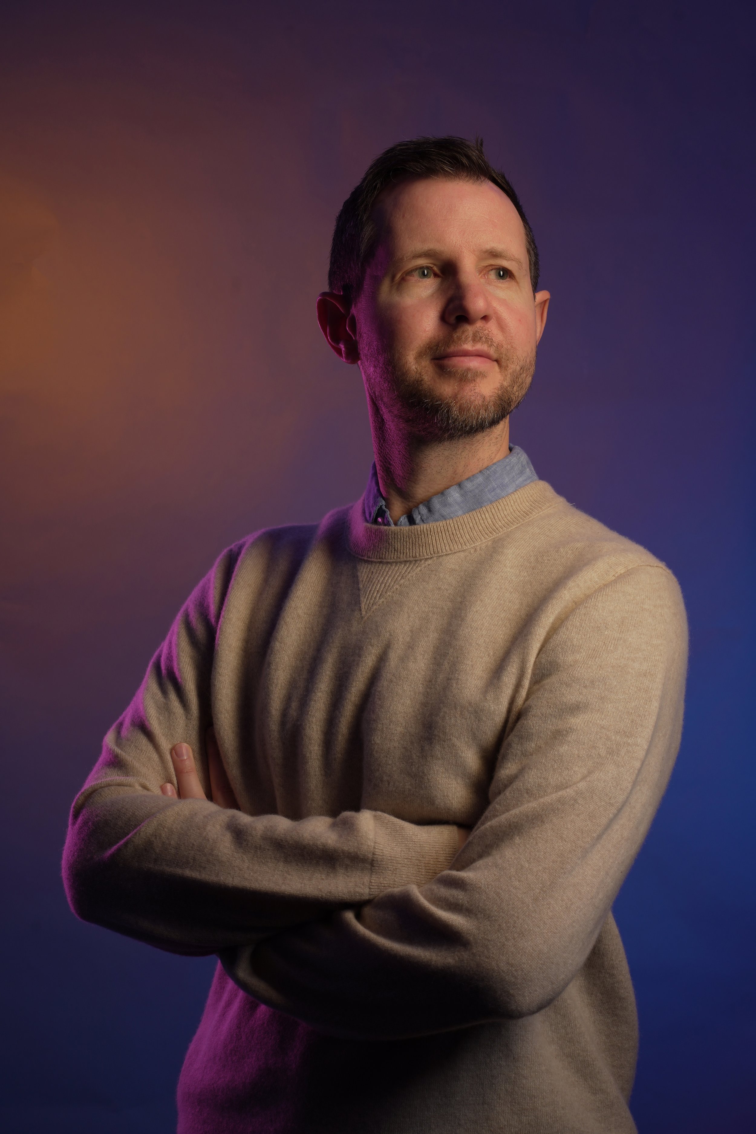 A man standing with his arms crossed, wearing a beige sweater over a gray collared shirt, looking to the right against a purple and orange gradient background.