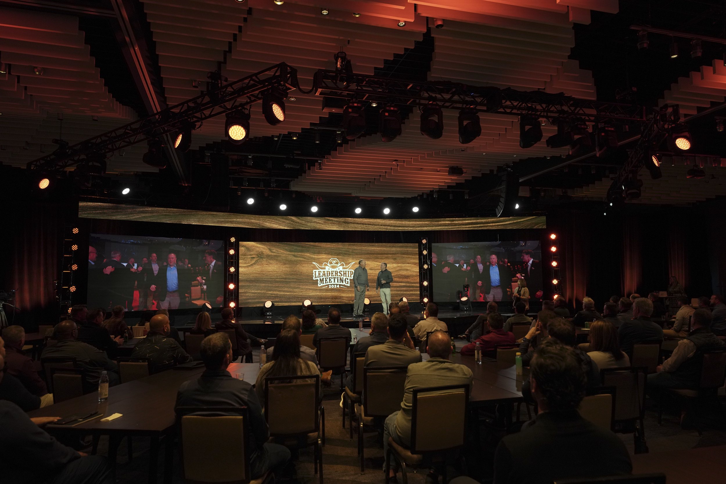 A large conference room with a stage and audience attending a Leadership Meeting 2024, with two people speaking on stage and a screen displaying the event logo.