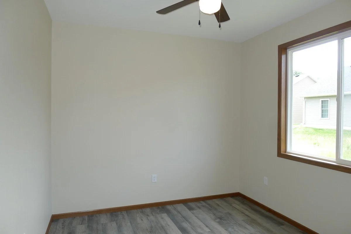 Empty room with beige walls, wood baseboards, grayish wood-look flooring, a ceiling fan with light, and a large window with a view of neighboring houses.