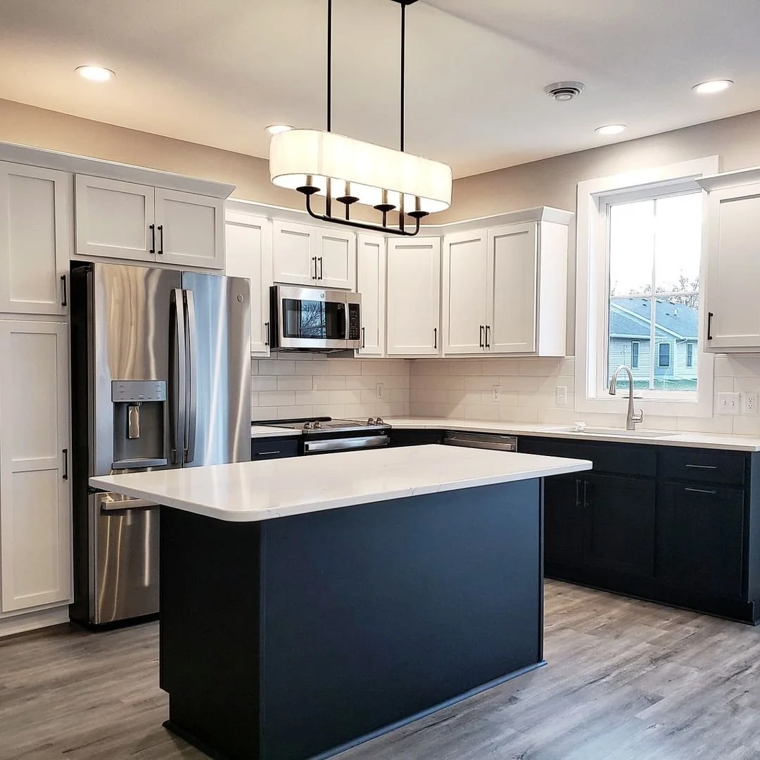 Modern kitchen with white and black cabinets, stainless steel refrigerator, microwave, and stove, island with white countertop, window above sink, pendant light fixture, and hardwood floor.