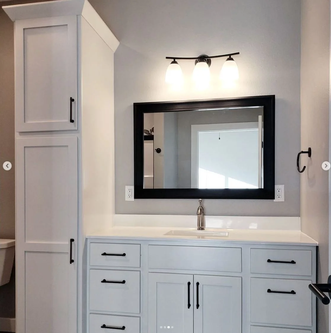 Bathroom vanity with white cabinets, black handles, a black-framed mirror, three-light fixture, faucet, and a wall towel hook.