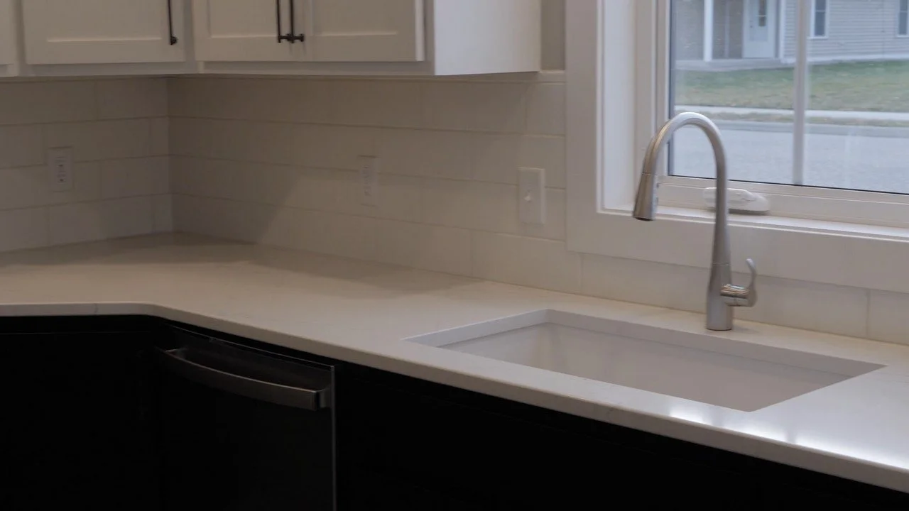 A modern kitchen with a white countertop, under-cabinet outlets, a stainless steel sink, and a high-arc faucet, next to a window with a view of outside houses and street.