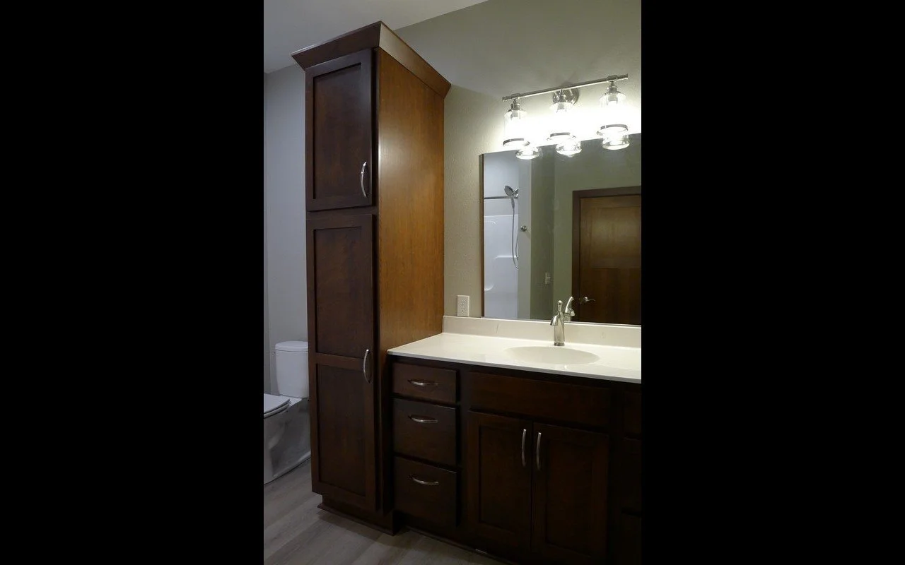 A bathroom with a dark wood vanity, a large mirror, a light fixture with multiple bulbs, and a toilet visible beside the vanity. There's a tall wooden cabinet next to the vanity and a door in the background.
