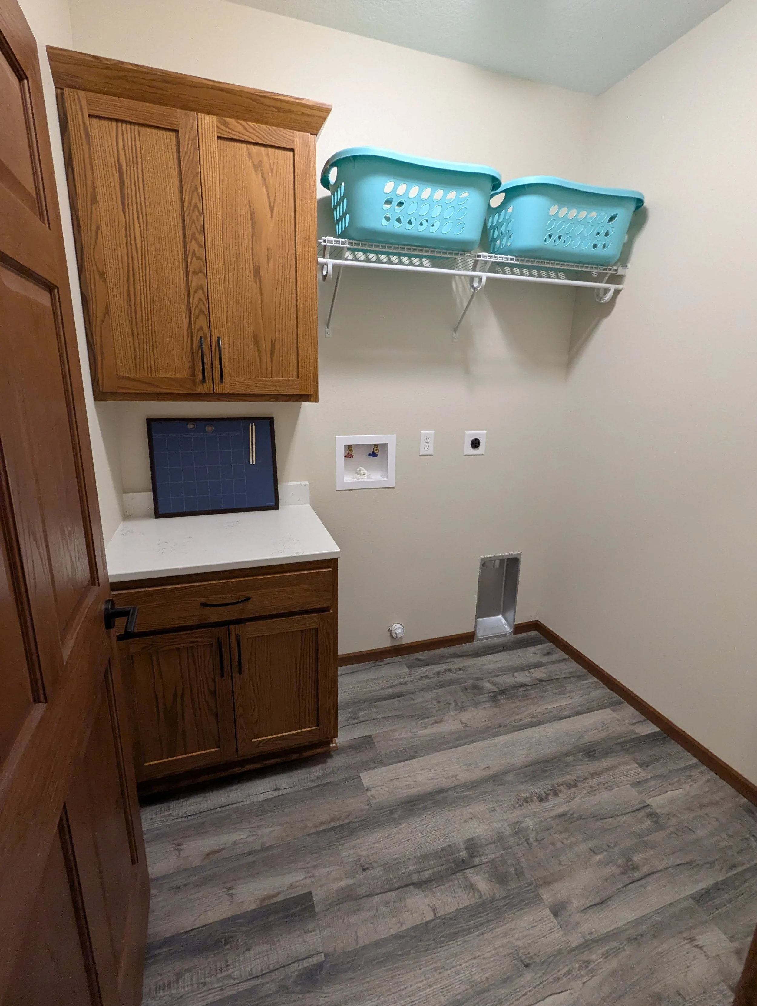 Empty laundry room with wooden cabinets, wire shelf with two teal laundry baskets, and a gray wood-look floor.