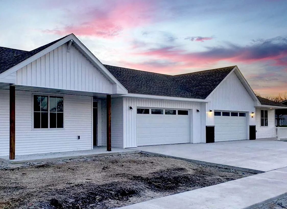 New white house with two garage doors, siding, and a front porch with wooden posts at sunset.