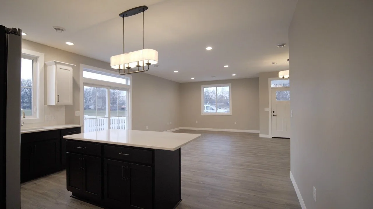 Empty modern kitchen and living area with large windows, island with dark cabinets and a white countertop, light gray walls, and wood flooring.