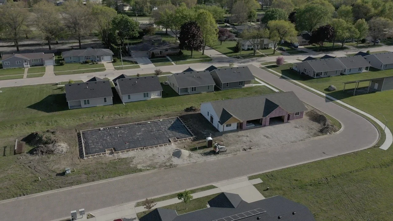 Aerial view of a residential neighborhood with houses, trees, and lawn areas, showing a house under construction and a foundation being prepared.