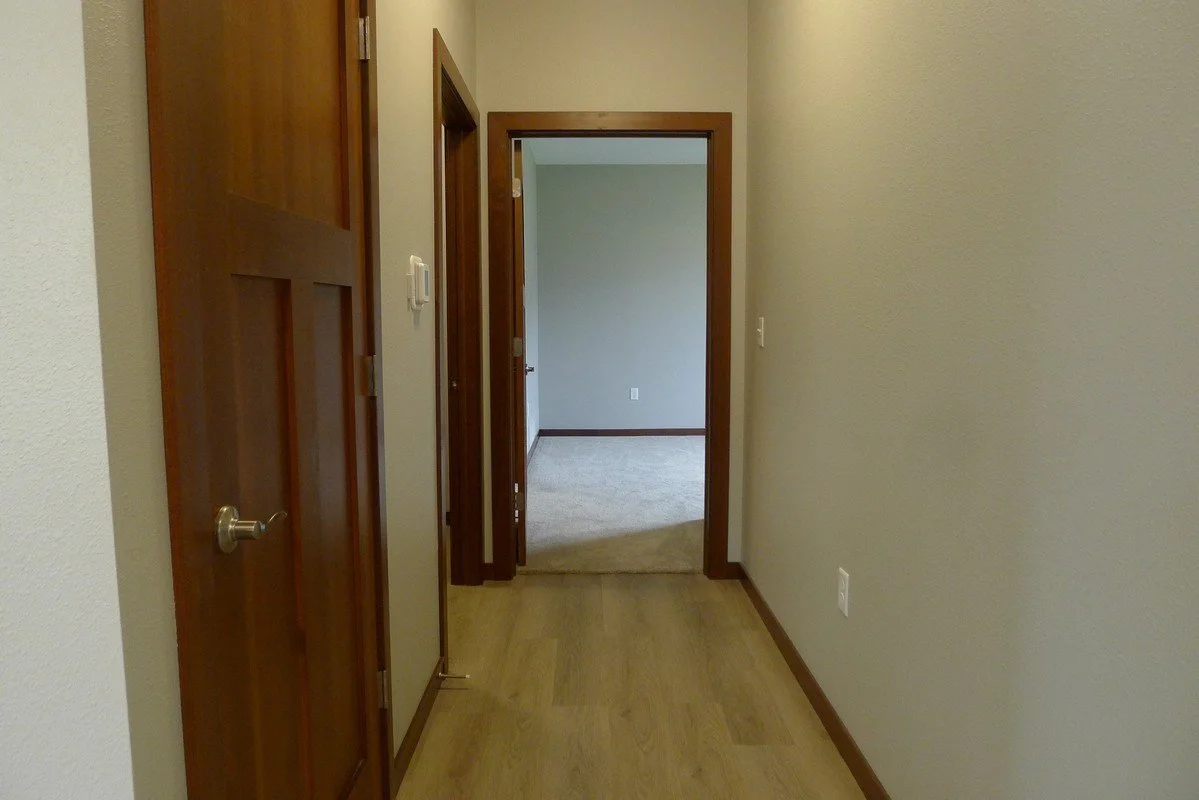 A hallway inside a house with wooden floors and doors, leading to a room with beige carpet and light green walls.