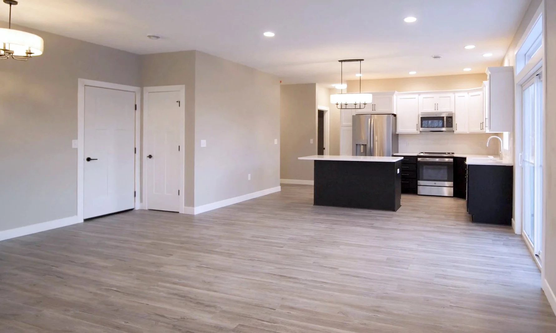 Empty modern kitchen and living area with light wood flooring, white and black cabinets, stainless steel appliances, and recessed ceiling lights.