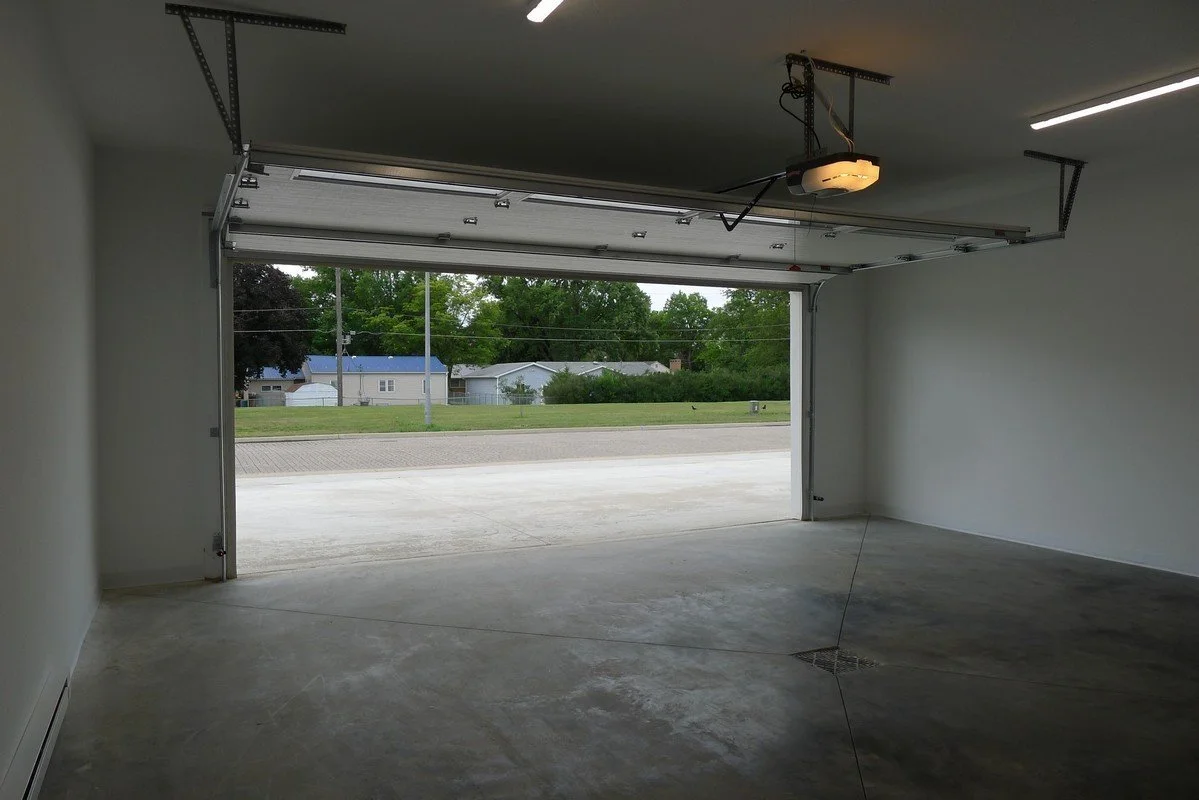 Empty garage with an open door revealing a suburban street with houses and trees outside.