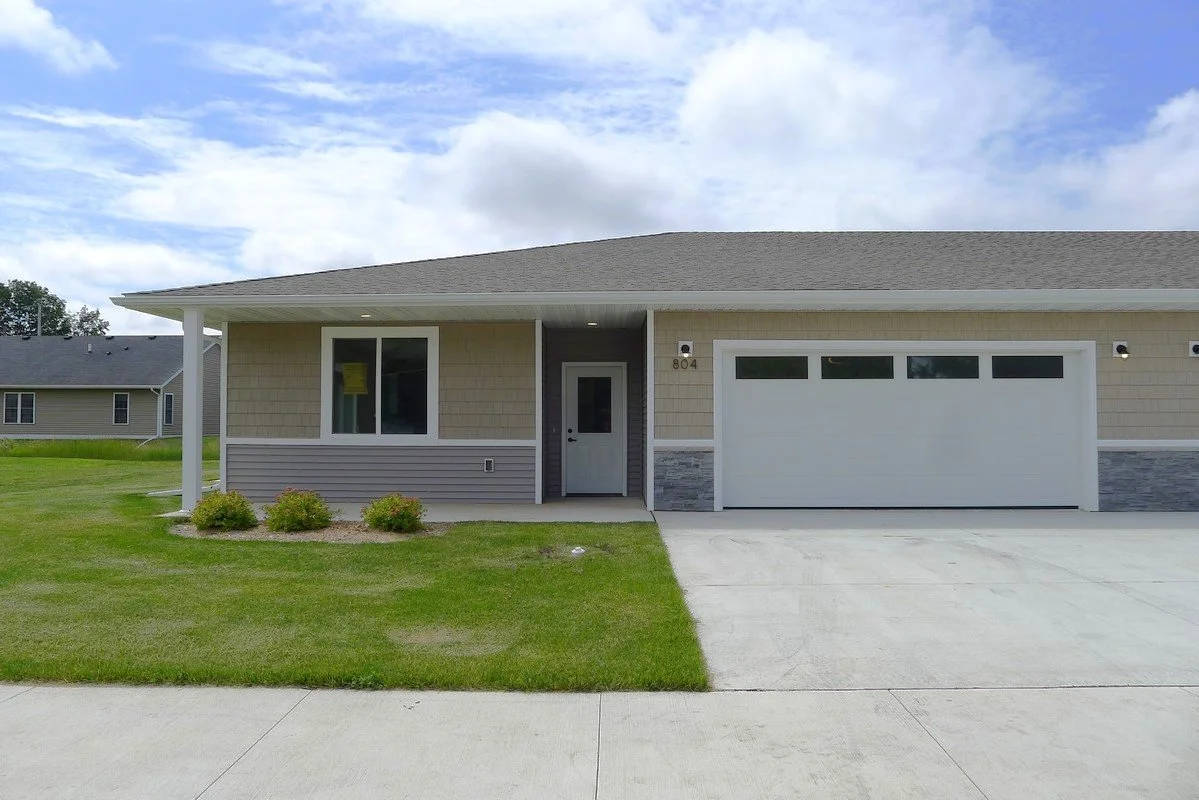 Front view of a modern single-story house with a lawn, driveway, and garage. The house has beige siding, white trim, and a gray stone accent at the entrance. The sky is partly cloudy.
