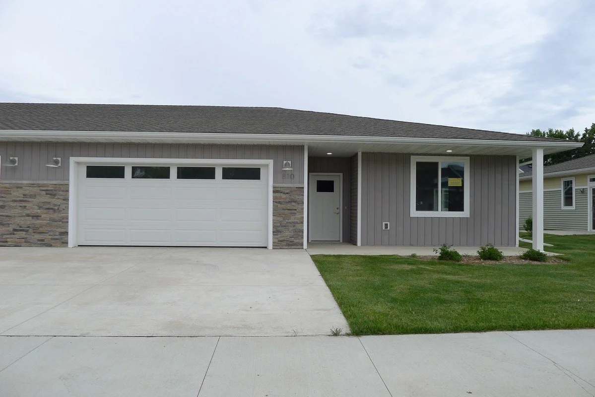 A modern single-story house with gray siding, a stone accent wall, a white garage door with windows, a small front door, and a window to the right. There is a concrete driveway, a small lawn with shrubs, and a porch with a white support column.
