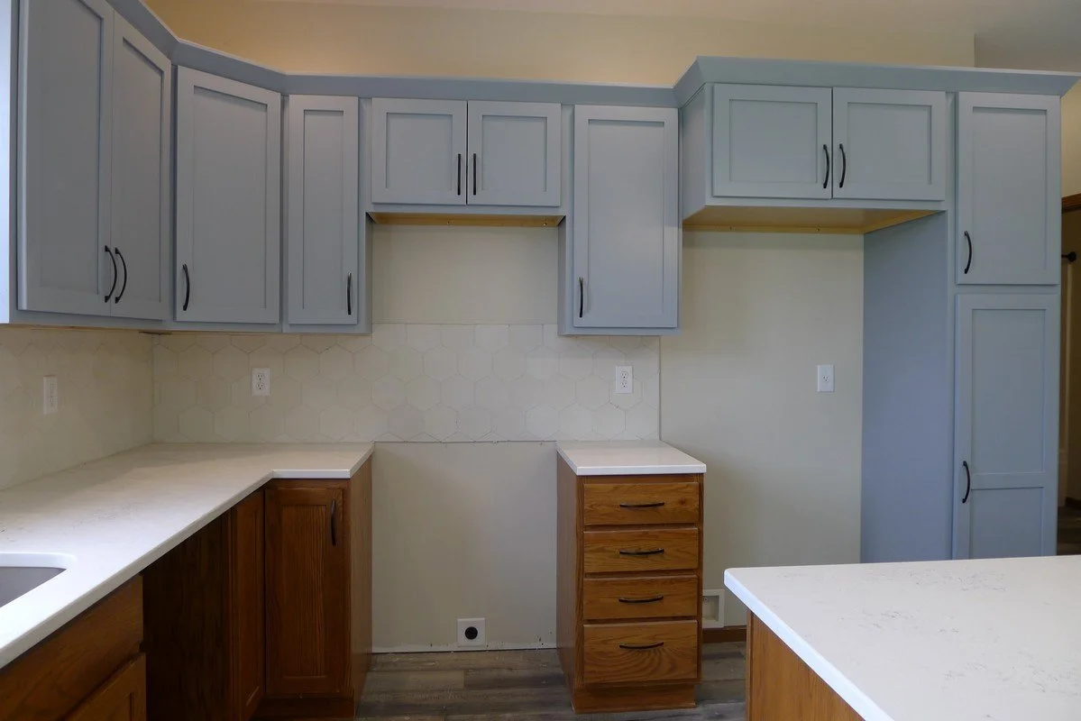 Empty kitchen with light blue upper cabinets, natural wood lower cabinets, and white countertops, limited appliances visible.