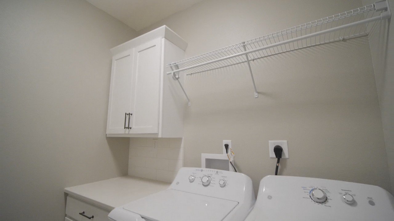 Laundry room with white washing machine and dryer, white cabinet, wire shelf, electrical outlets, and wall hook.
