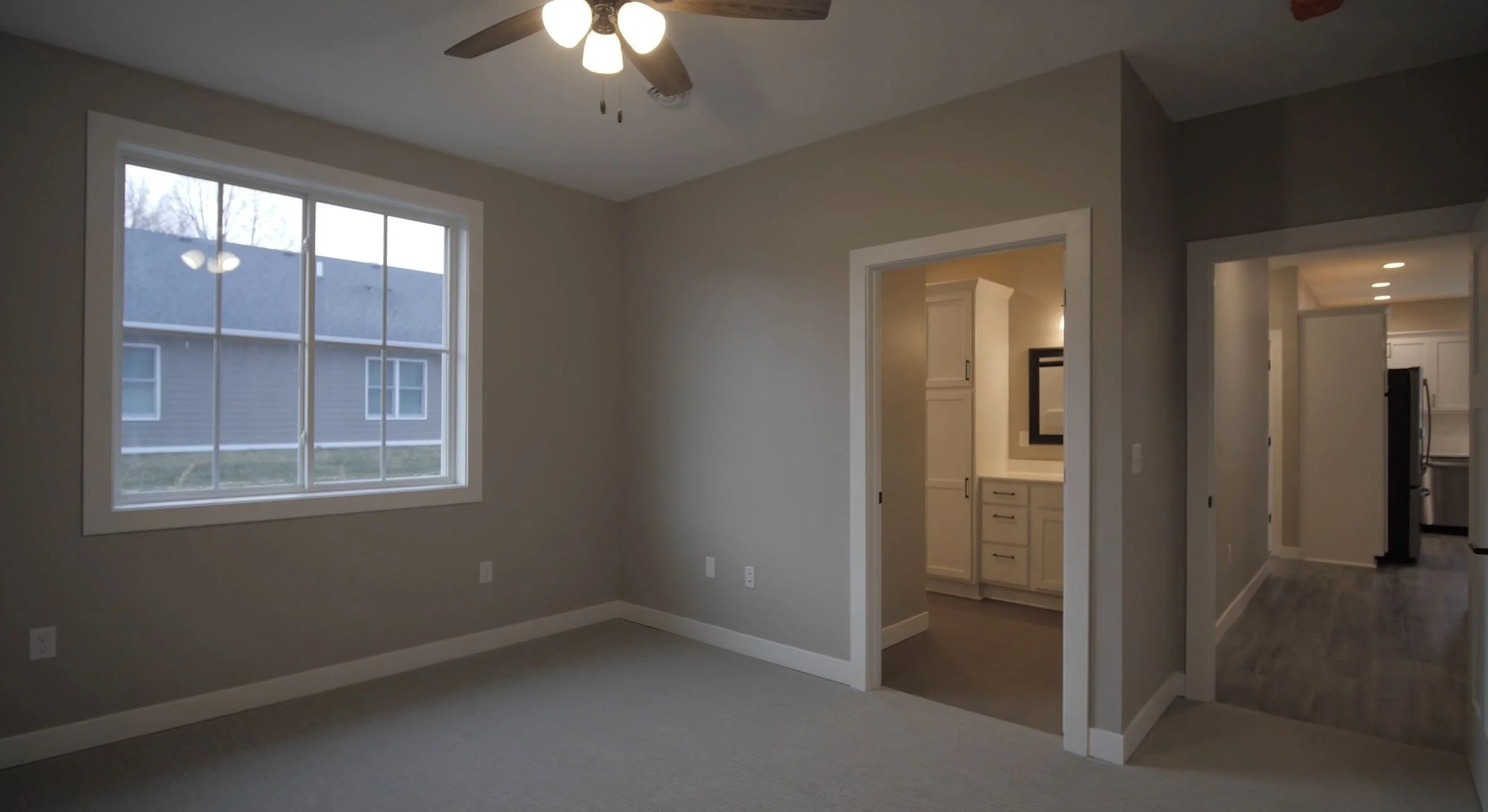 Interior view of an empty room with beige walls, a large window, ceiling fan, and adjacent hallway leading to kitchen and bathroom.