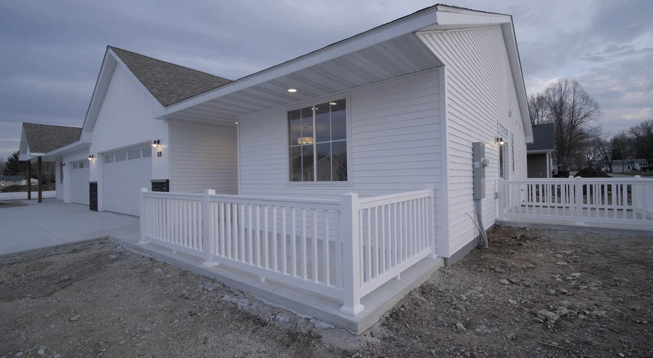 Newly built white house with a small porch, white railing, large window, and garage, set against a cloudy sky.