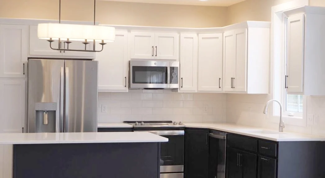 Modern kitchen with white upper cabinets, black lower cabinets, stainless steel refrigerator, microwave, and oven, white countertops, a window above the sink, and a contemporary chandelier.