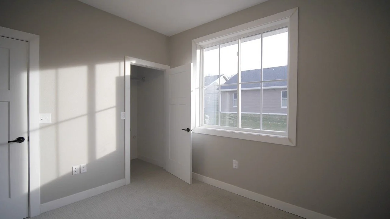 Empty bedroom with a large window, closet, and plain light-colored walls.
