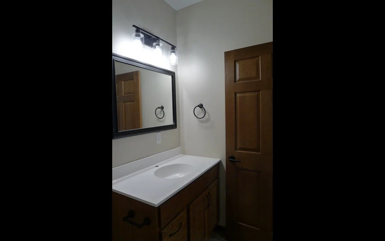 A small bathroom vanity with a white countertop, an integrated sink, a black framed mirror, and a wall-mounted towel ring. The cabinetry is wooden with black handles. There is a wooden door next to the vanity, and a three-bulb light fixture above the