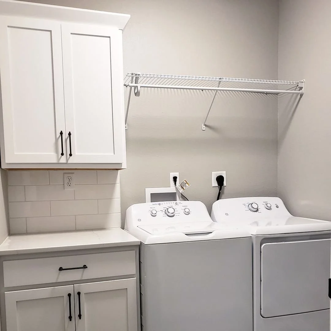A laundry room with white cabinets, a countertop, a washing machine, and a dryer. There is a wire shelf above and two electrical outlets behind the machines.