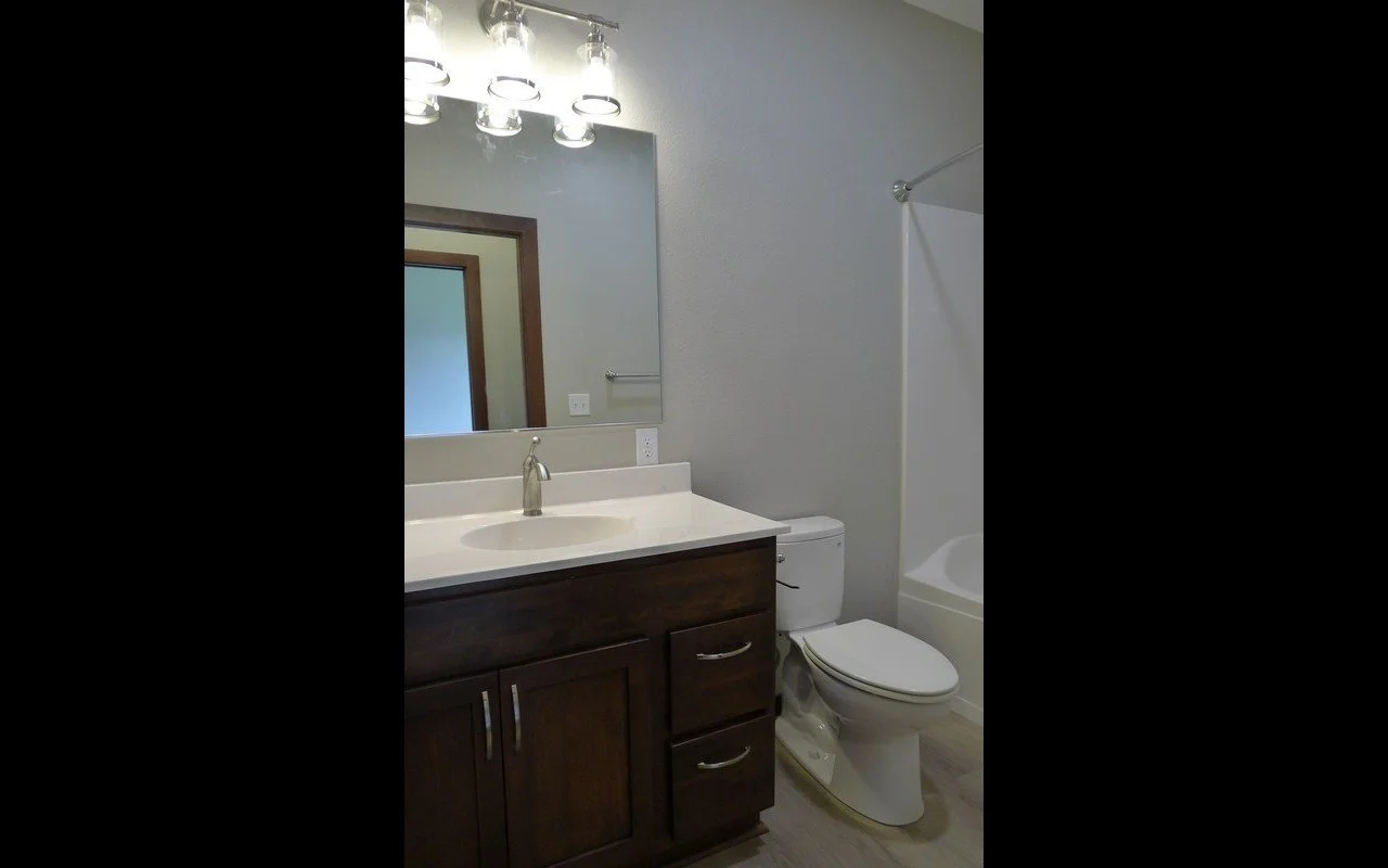Bathroom with dark wood vanity, white countertop, mirror with modern light fixture, toilet, and bathtub with shower curtain rod.