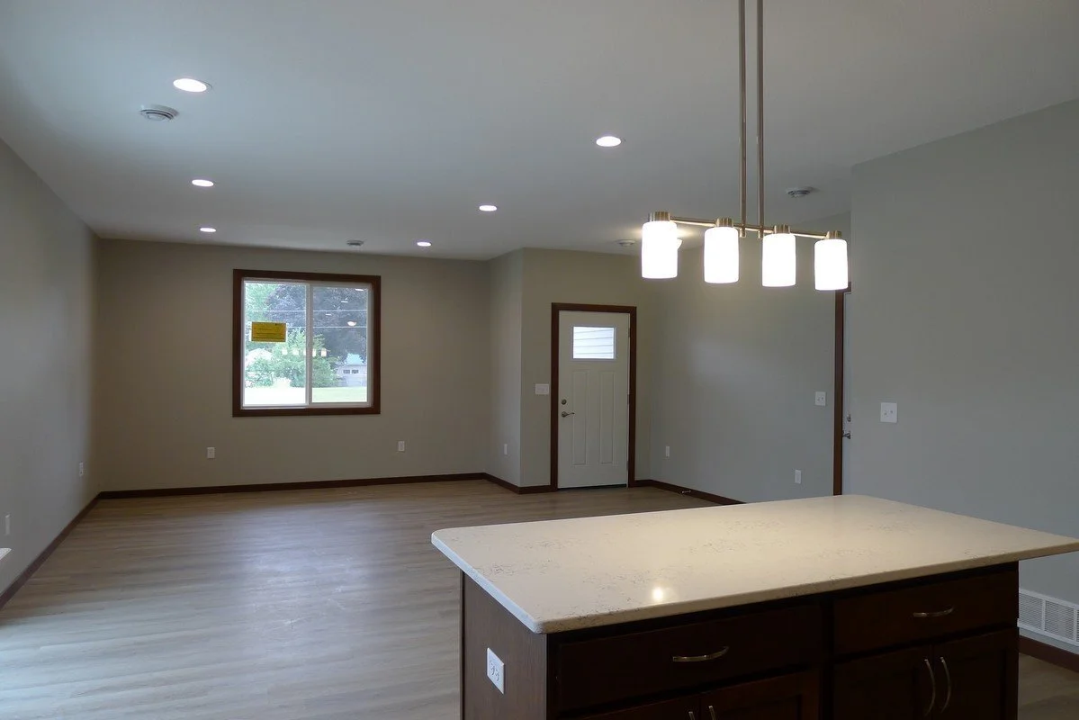 Empty living room with hardwood floors, a window, and a front door, connected to kitchen with island and modern lighting.