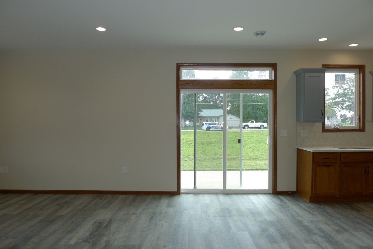 Empty living space with a sliding glass door, a window, wooden cabinets, and a view of a green lawn and parked cars outside.