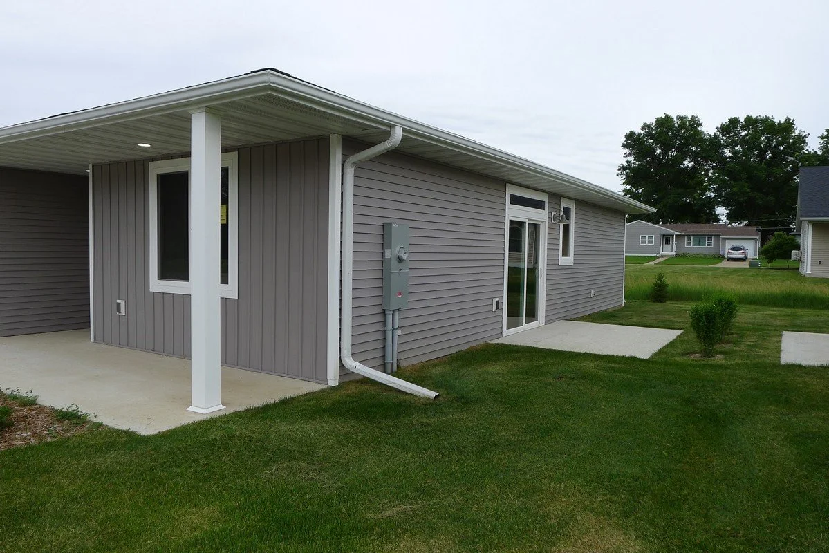 Backyard of a house with gray siding, sliding glass door, small window, utility meter, and a covered patio area with white support posts, grassy lawn, and concrete slabs leading to the door.