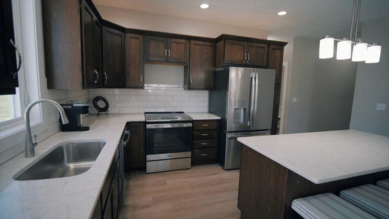 Kitchen with dark wood cabinets, white marble countertops, stainless steel refrigerator and stove, white subway tile backsplash, window above sink, and modern lighting fixture.