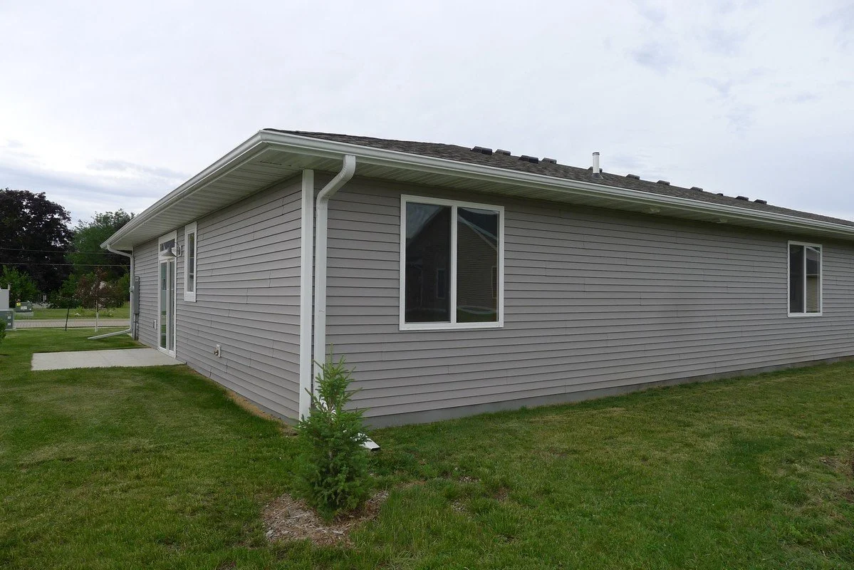 Side view of a house with gray siding, white trim, and a small front yard with a young tree, under an overcast sky.