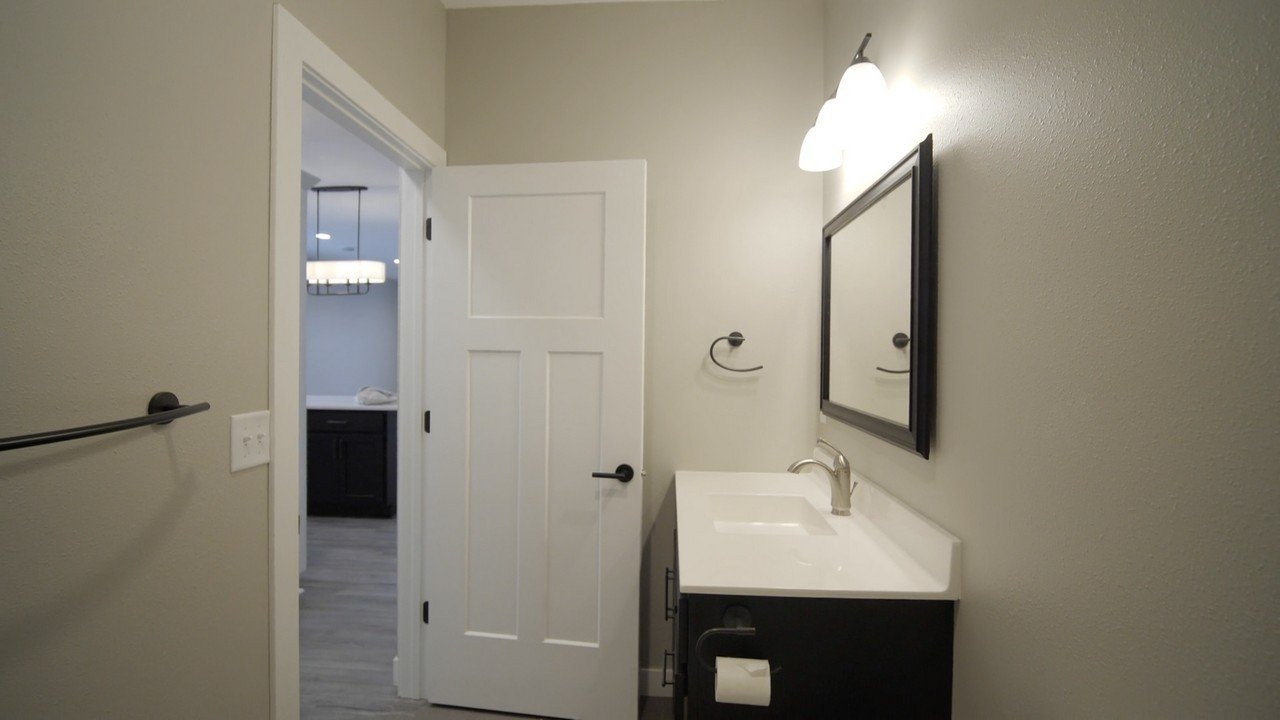 Bathroom with a vanity mirror, sink, and a wall light fixture, with a towel bar and an open door leading to a room with a chandelier.