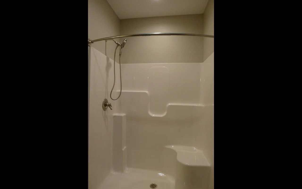 Empty shower with white walls, metal shower rod, handheld showerhead, and built-in seat.