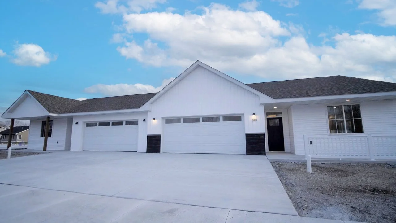 A modern white house with three garage doors, a black front door, and a dark shingled roof, under a partly cloudy sky.