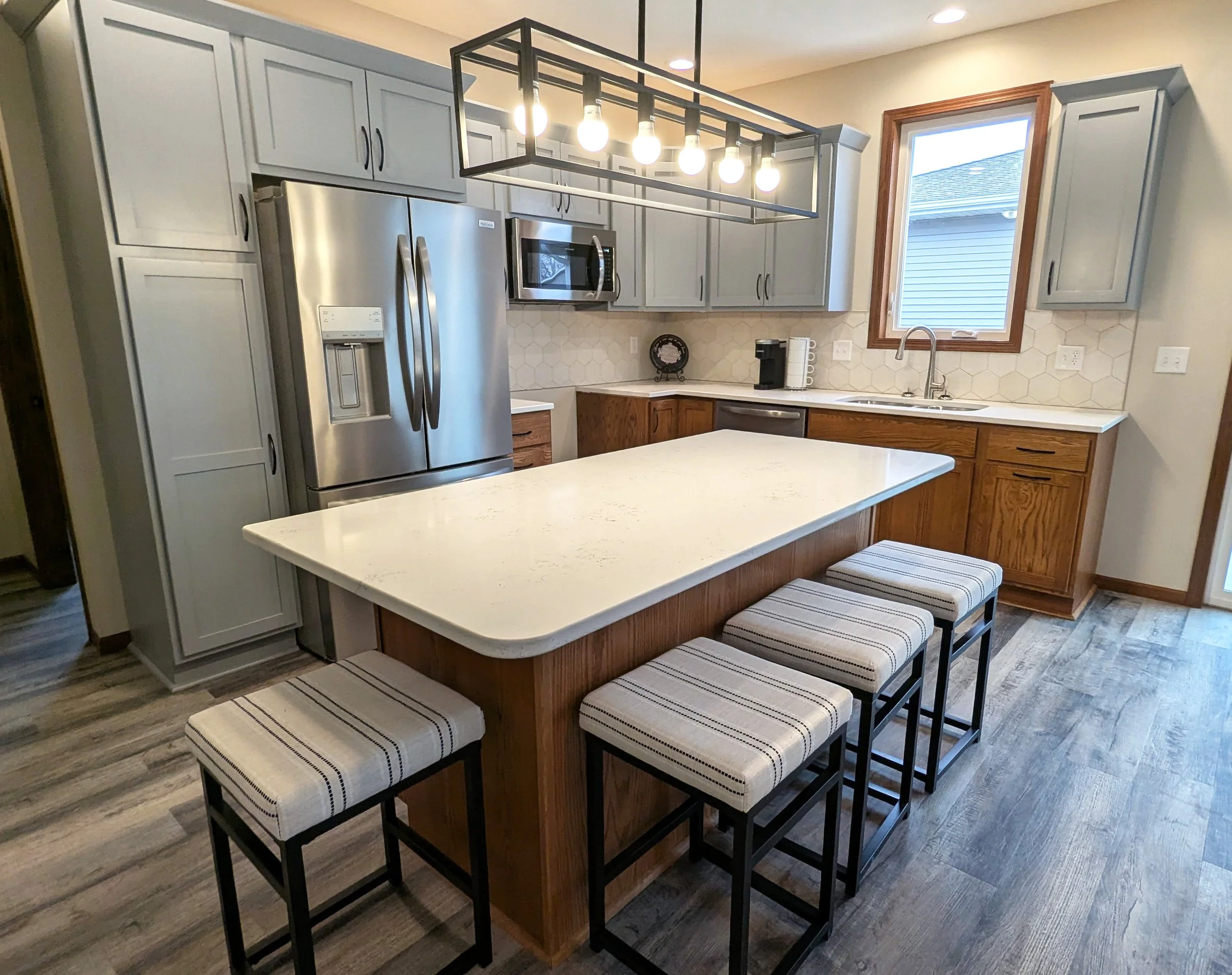 Modern kitchen with gray upper cabinets, wooden lower cabinets, stainless steel refrigerator, microwave, and dishwasher. An island with a white countertop has four striped upholstered barstools. There is a window above the sink, and a black hanging l
