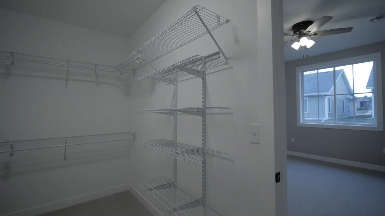 Empty walk-in closet with white wire shelves and a ceiling fan in an adjacent room with a large window.