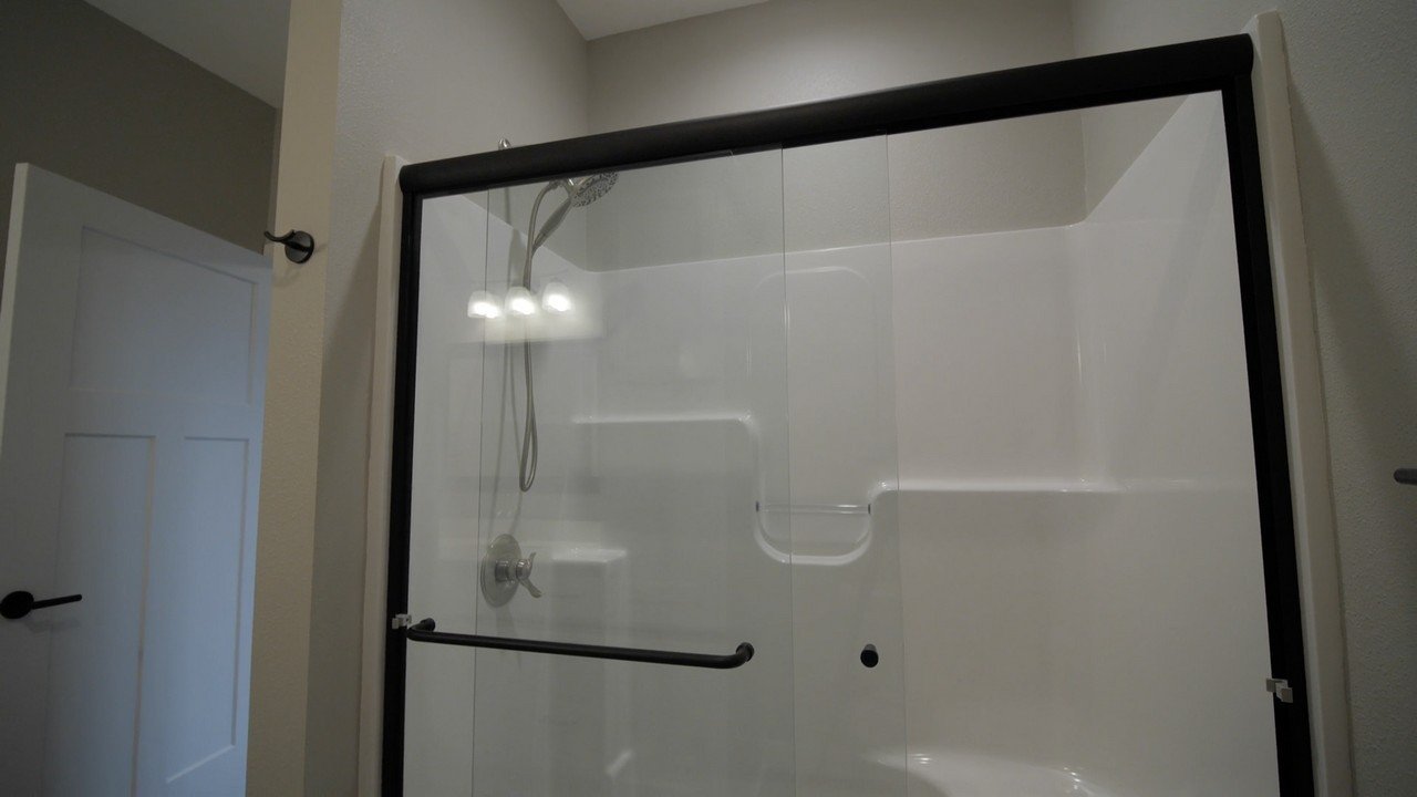 A walk-in shower with clear glass doors, a handheld showerhead, and built-in shelves, in a bathroom with a white door and beige walls.