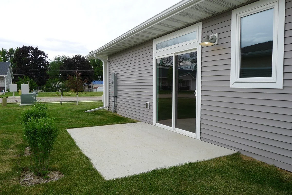 Backyard patio area with a concrete pad next to a gray house with vinyl siding, sliding glass door, and a small window. Outdoor light fixture on the wall and green grass with small bushes nearby.