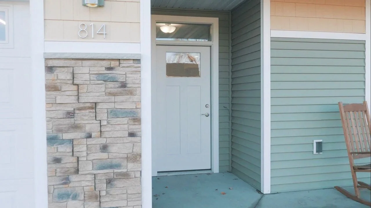 Close-up of a house entrance with a gray door, stone wall siding, and house number 814 above the door. Part of a wooden rocking chair is visible on the right.
