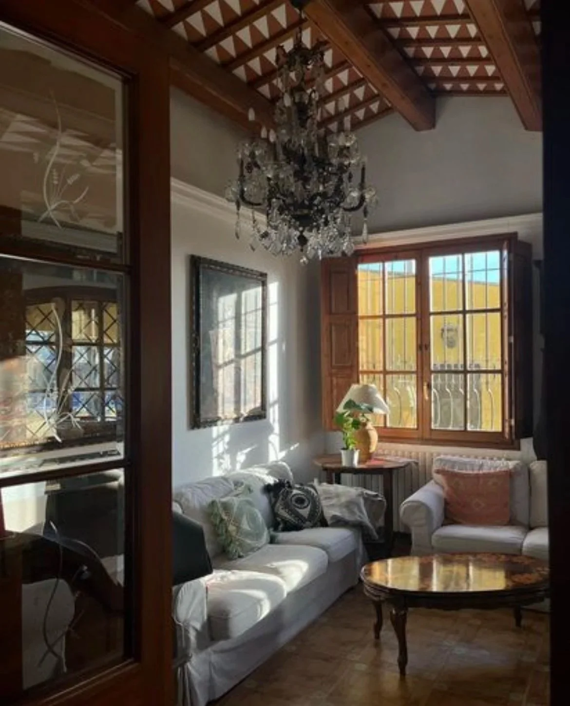 Living room with white sofa, wooden coffee table, window with sunlight, potted plant, and chandelier.