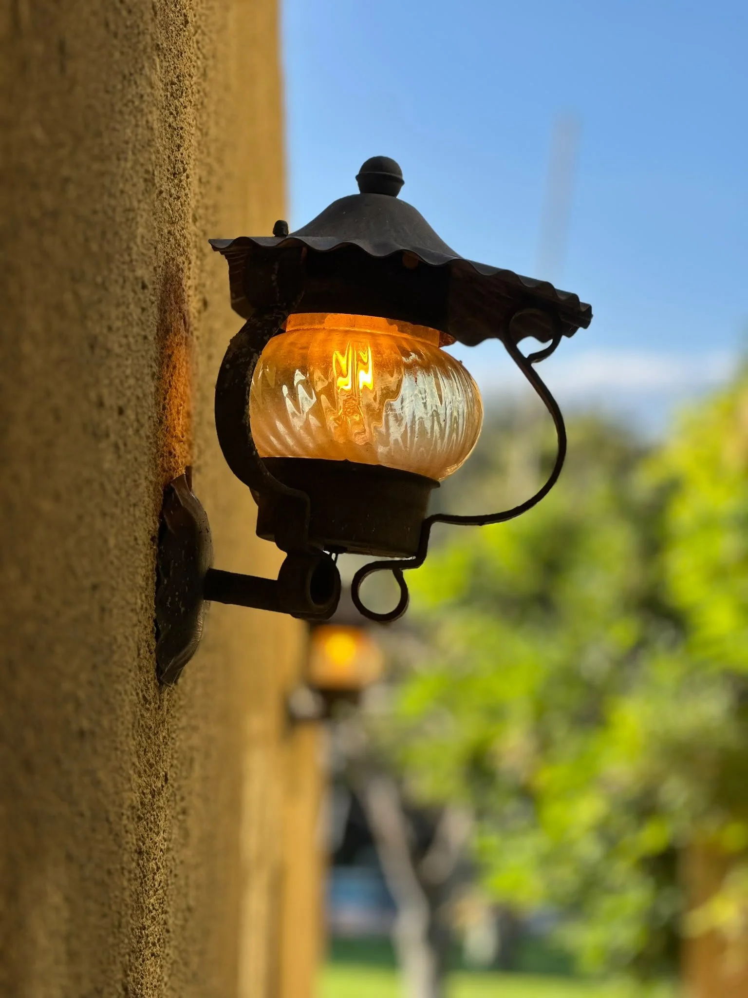 Close-up of an outdoor vintage-style lantern with a glowing light, mounted on a textured beige wall with trees and blue sky in the background.