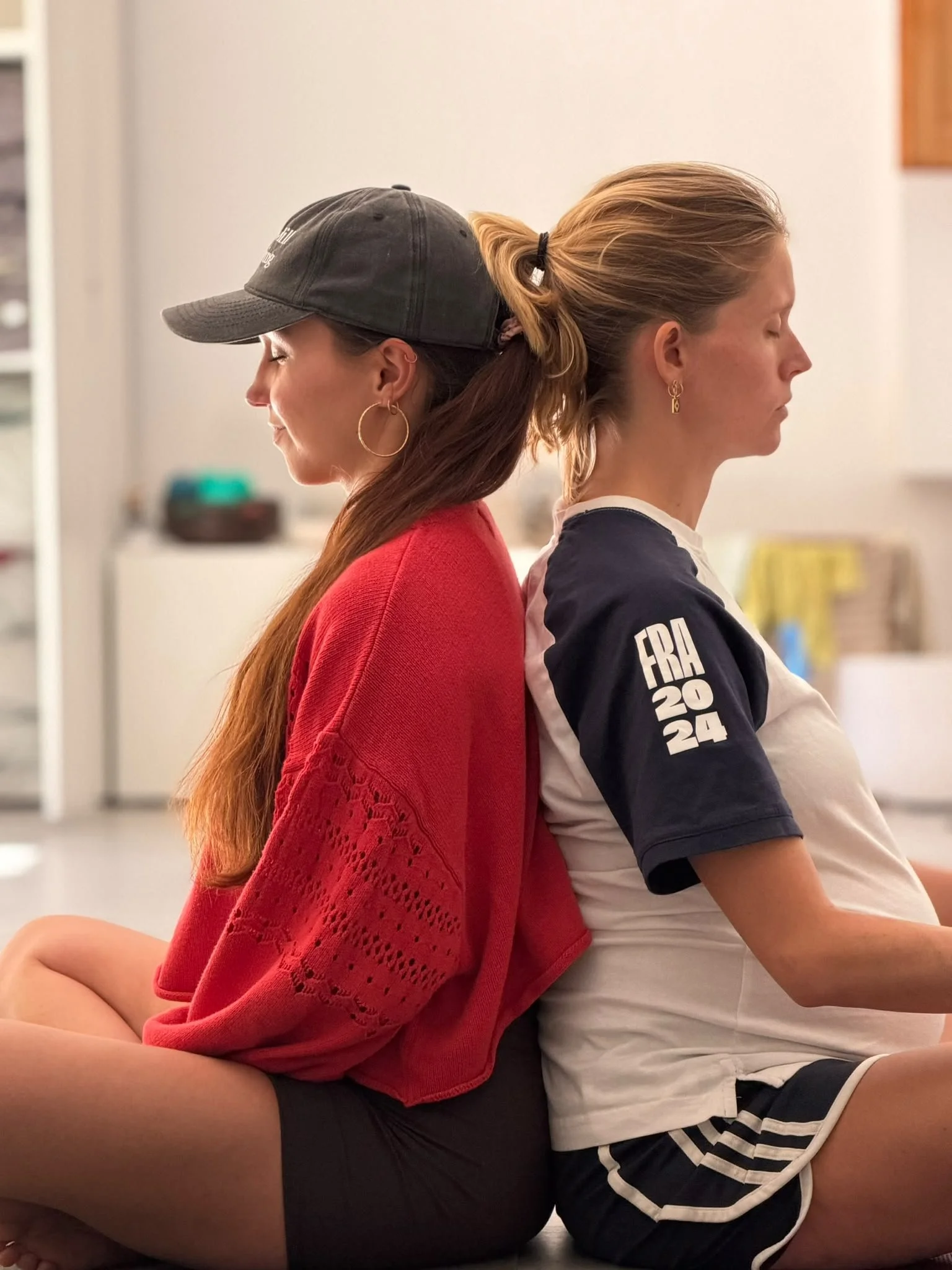 Two women sit with their backs against each other and eyes closed in a calm, indoor setting.
