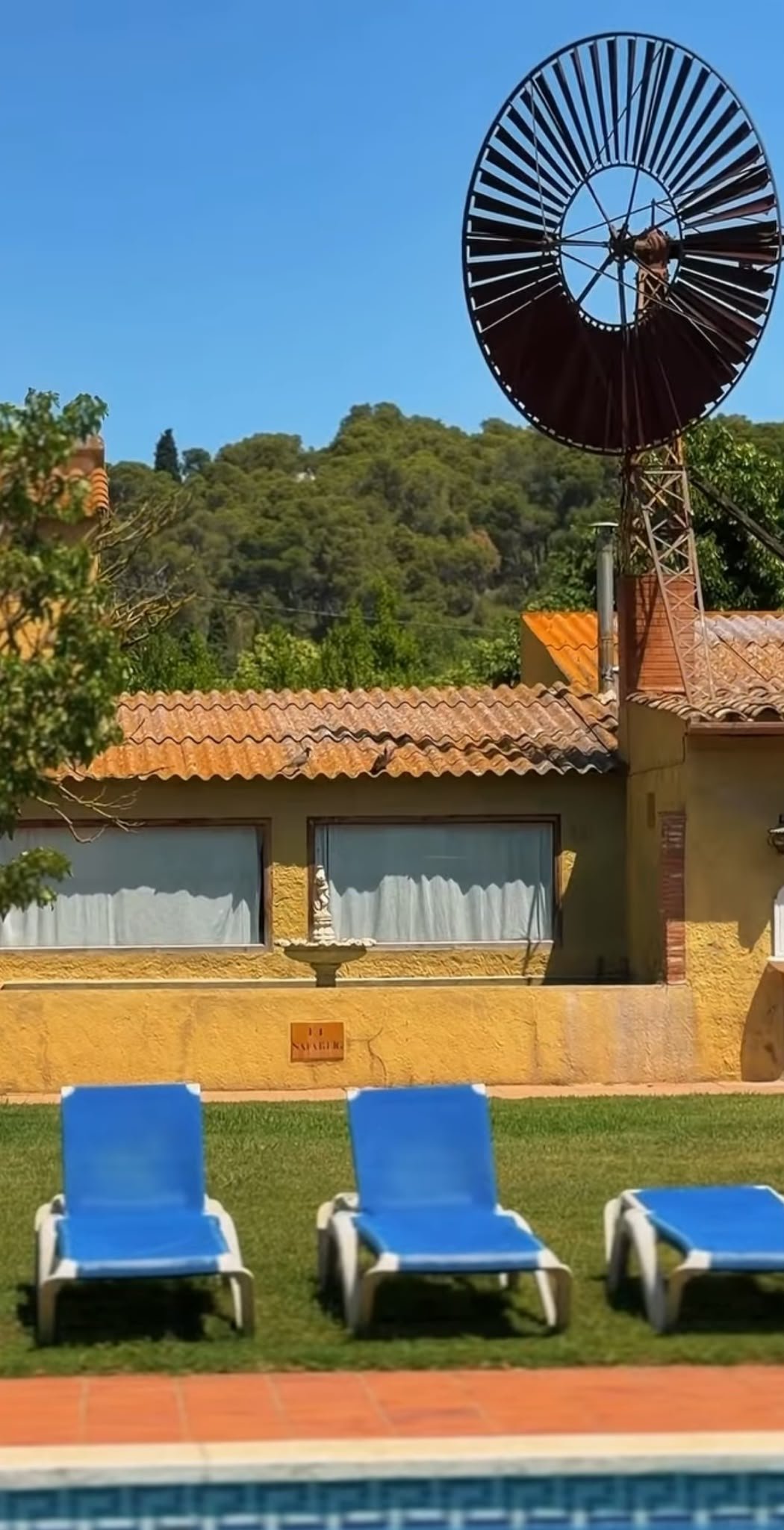 Three blue pool chairs on a grassy area in front of a yellow building with a tiled roof and a window with curtains, with a small statue on a pedestal and trees behind, and a large windmill structure on the roof.