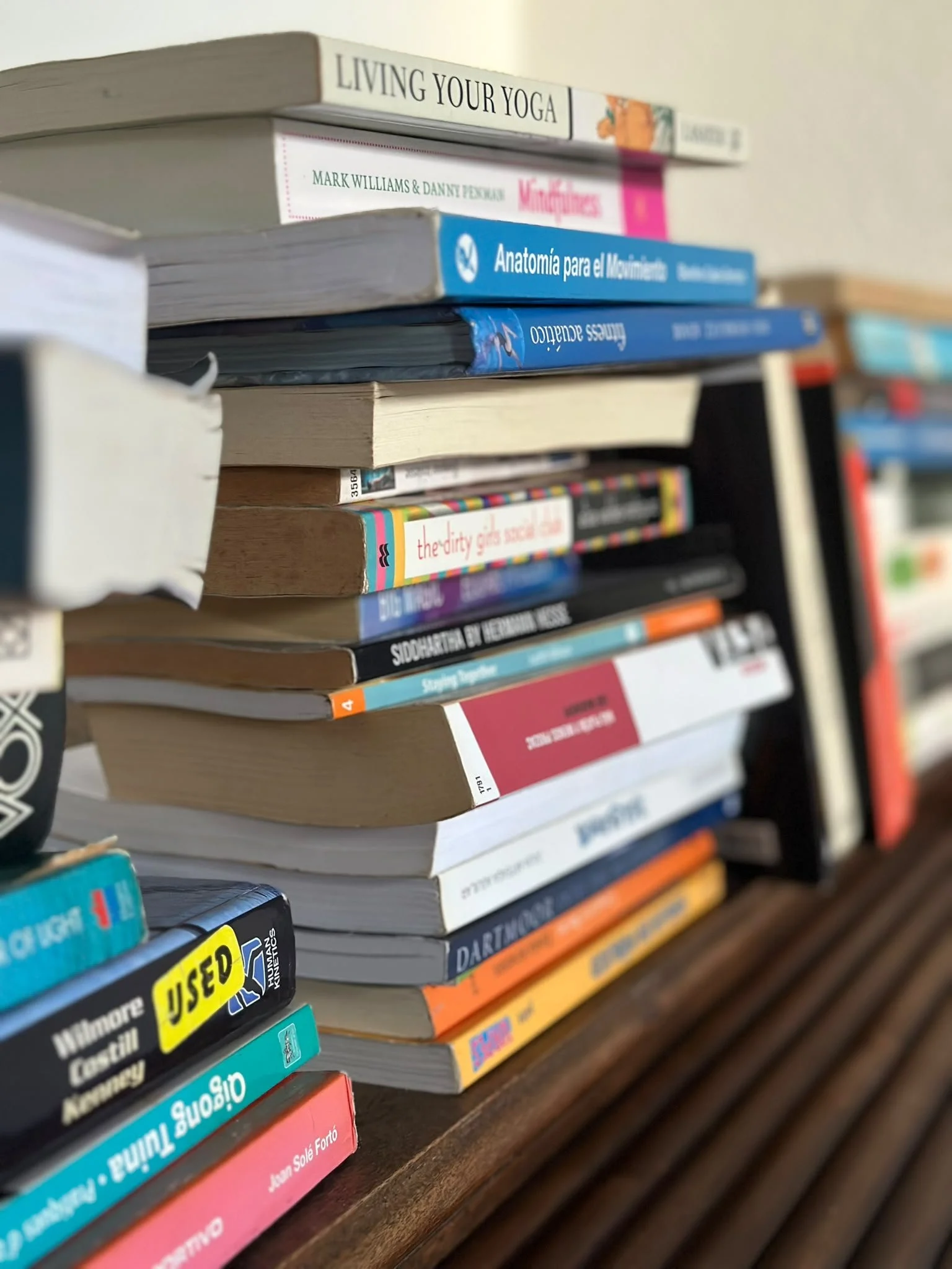 Close-up of a stack of books and magazines on a wooden surface, with titles related to yoga, anatomy, and other topics, some worn and used.