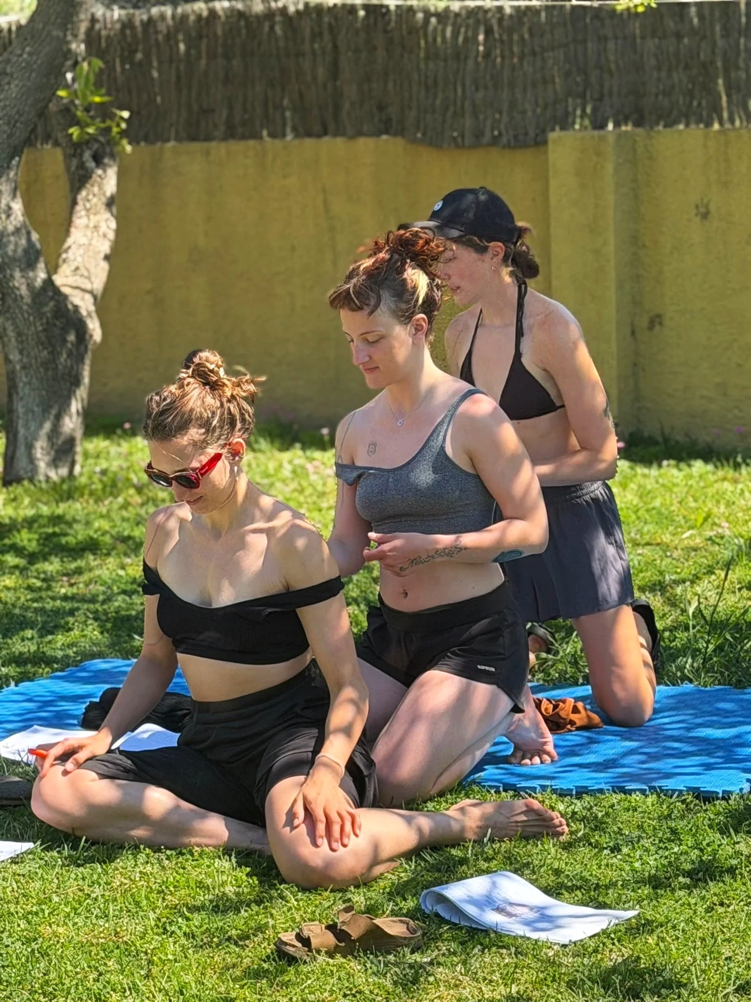 Three women practicing yoga outdoors on a grassy area on a sunny day, with one woman sitting cross-legged and the other two kneeling behind her. They are on yoga mats. There are trees, a wooden fence, and a yellow wall in the background.