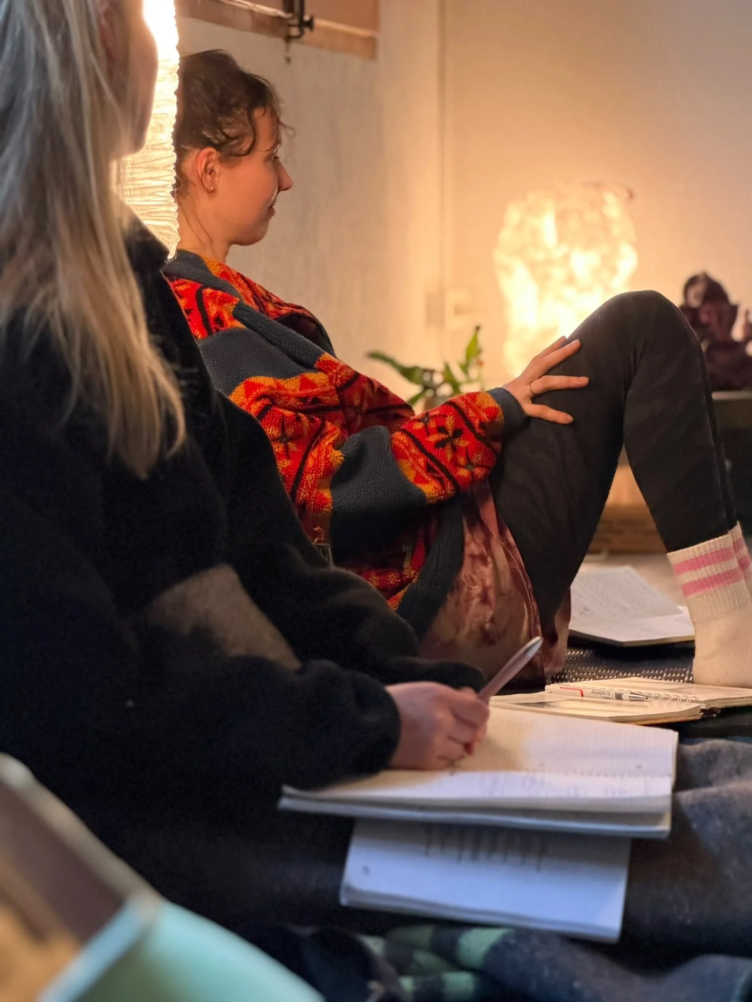 Two women sitting at a table, one reading a book and the other holding a phone in a cozy room with warm lighting.