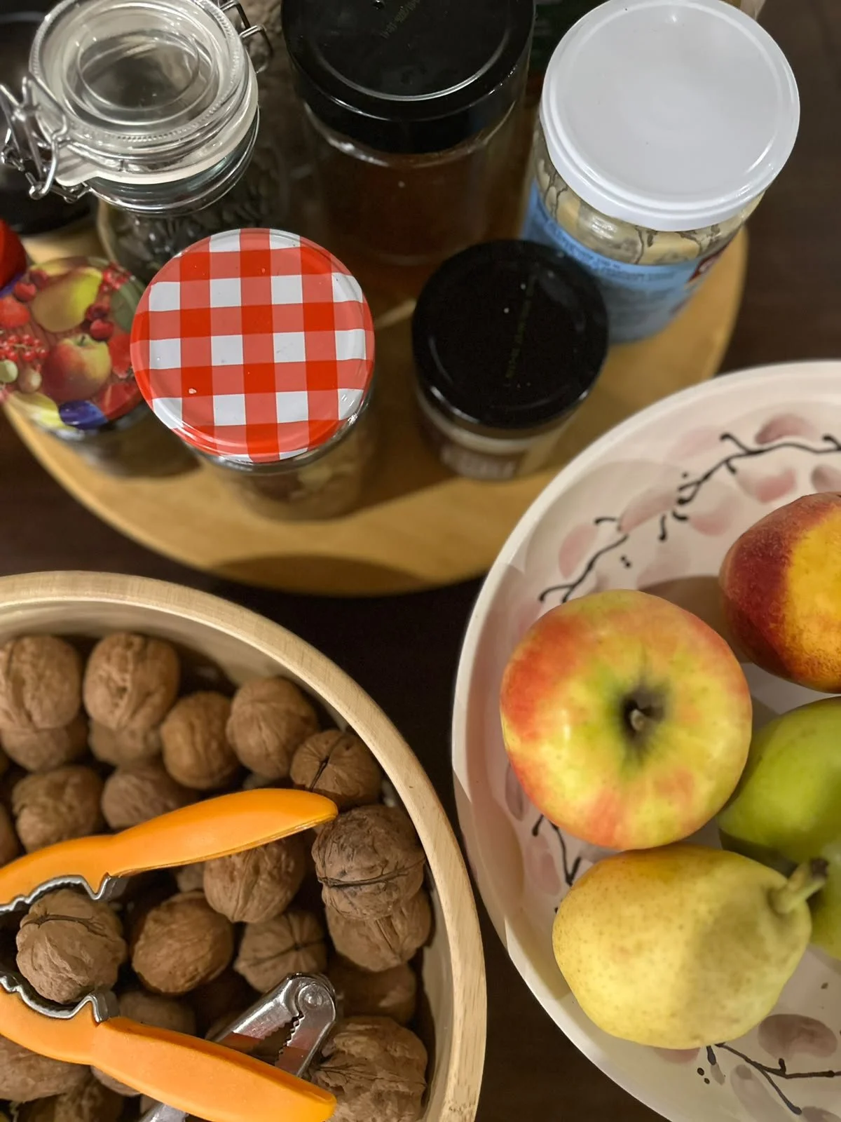 Top view of a round wooden tray with spice jars with black, clear, and checkered red and white lids, next to a bowl of apples and a bowl of walnuts. The apples are red and yellow, and the walnuts are in a round container with orange tongs.