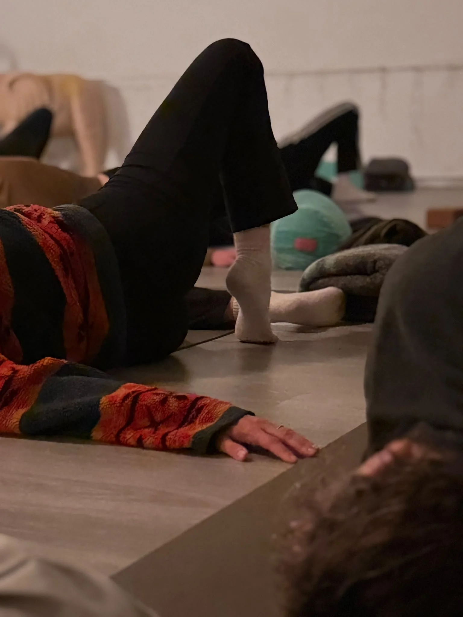 People practicing yoga indoors, lying on their backs with legs lifted, in a relaxed setting with mats and personal belongings nearby.